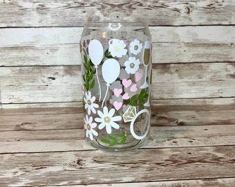 May include: A clear glass can with a white, pink and gold design. The design features flowers, hearts, balloons and a ring with a diamond.
