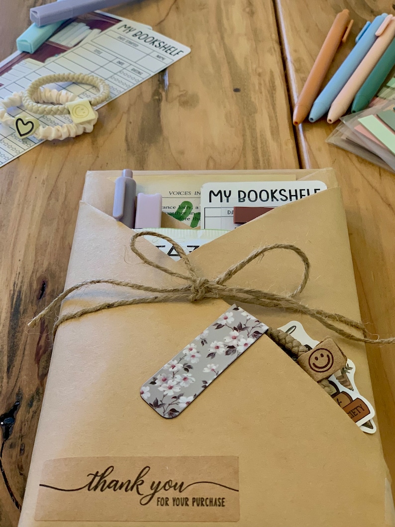 May include: A brown paper package tied with twine and decorated with a floral bookmark, a smiley face sticker, and a "thank you" sticker. The package contains a notebook with the text "My Bookshelf" on the cover.