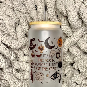 May include: A frosted glass tumbler with a bamboo lid and a clear straw. The tumbler has a Halloween-themed design with a black and white illustration of a witch's hat, a crescent moon, a pumpkin, a skull, a potion bottle, a book, a snake, a cauldron, a feather, a candle, and an eye. The text on the tumbler reads "It's the most wonderful time of the year."