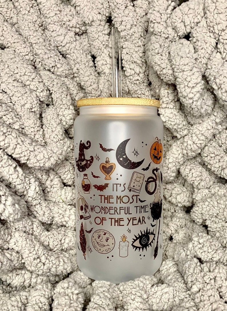 May include: A frosted glass tumbler with a bamboo lid and a clear straw. The tumbler has a Halloween-themed design with a black and white illustration of a witch's hat, a crescent moon, a pumpkin, a skull, a potion bottle, a book, a snake, a cauldron, a feather, a candle, and an eye. The text on the tumbler reads "It's the most wonderful time of the year."