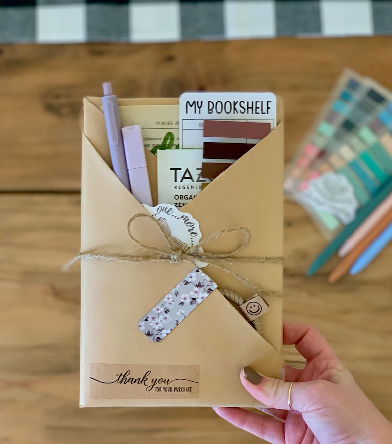 May include: A brown paper envelope tied with string and a "thank you" sticker. Inside the envelope is a notebook, pens, and a card that reads "My Bookshelf".