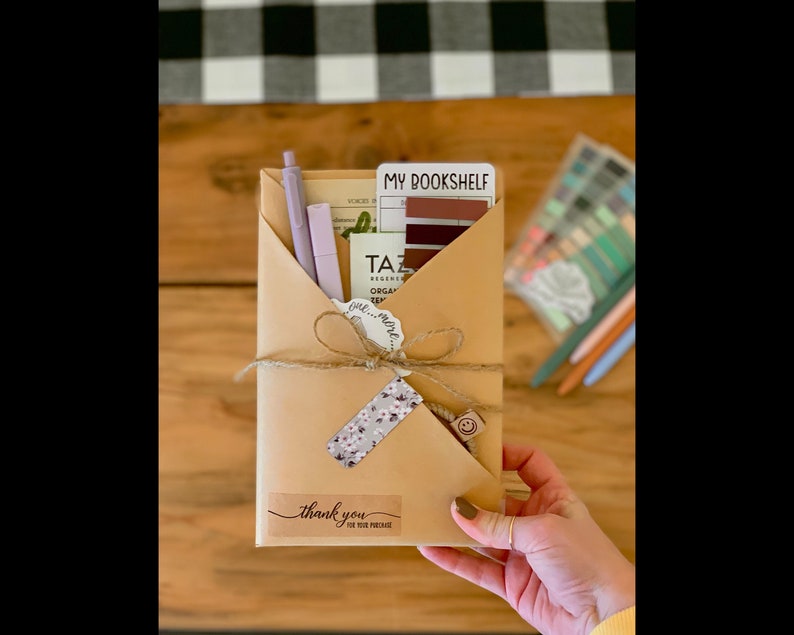 May include: A brown paper envelope with a thank you note, a small floral tag, and a sticker that says "My Bookshelf". The envelope is tied with twine and has a small wooden stamp with a smiley face on it. There are also two purple pens and a small card with the text "Tazas" on it.