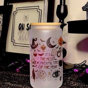 May include: A frosted glass tumbler with a bamboo lid and a stainless steel straw. The tumbler has a Halloween-themed design with a variety of spooky icons, including a witch's hat, a crescent moon, a skull, a cauldron, and a bat. The text on the tumbler reads "It's the most wonderful time of the year."