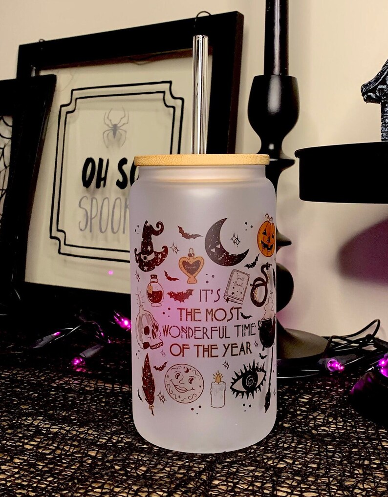 May include: A frosted glass tumbler with a bamboo lid and a stainless steel straw. The tumbler has a Halloween-themed design with a variety of spooky icons, including a witch's hat, a crescent moon, a skull, a cauldron, and a bat. The text on the tumbler reads "It's the most wonderful time of the year."
