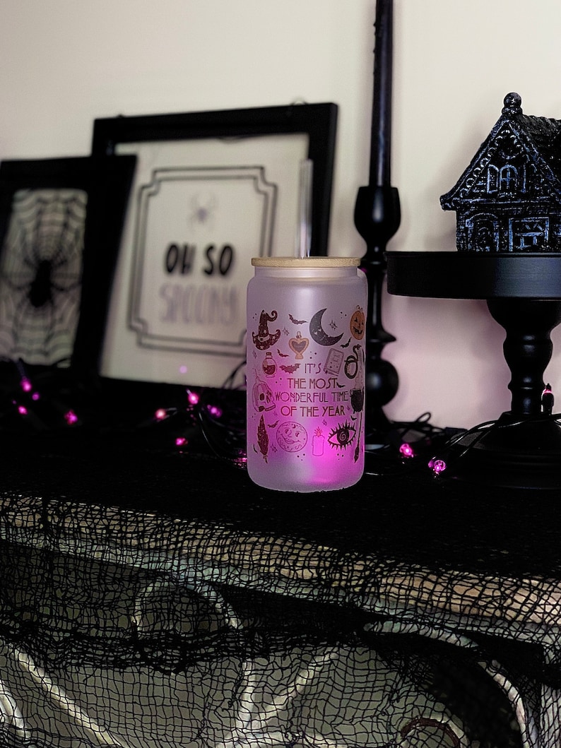 May include: A frosted glass jar with a wooden lid and a pink glow. The jar has a Halloween theme with illustrations of bats, a witch's hat, a crescent moon, a skull, an eye, and a potion bottle. The text on the jar reads "It's the most wonderful time of the year".