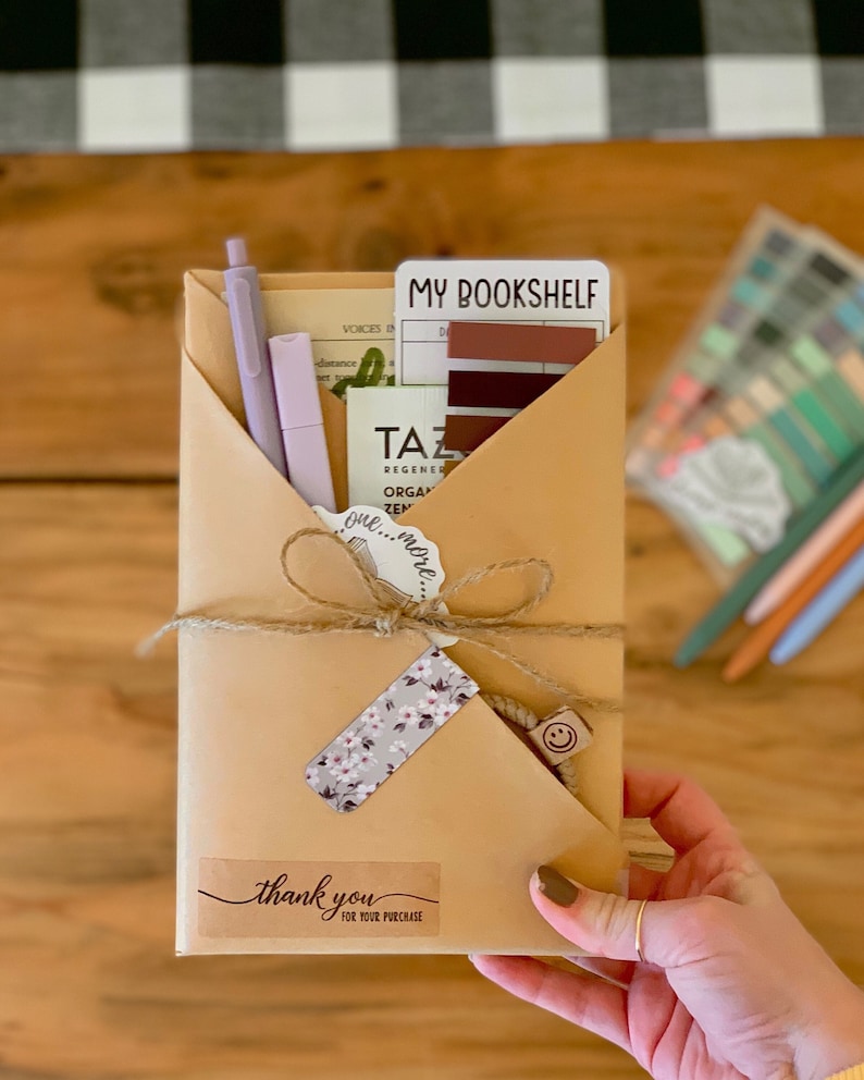May include: A brown paper envelope with a thank you sticker and a floral sticker. The envelope is tied with twine and has a small wooden smiley face charm attached. The envelope contains a variety of items, including pens, stickers, and a card that says "My Bookshelf".