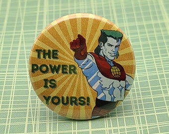 May include: Round button with a cartoon image of a superhero with green hair, blue skin, and a red cape. The button has the text "THE POWER IS YOURS!" against a yellow and orange sunburst background.