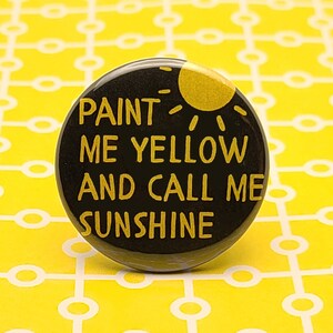 以下が含まれることがあります： 黄色い文字と黄色い太陽のグラフィックが描かれた黒いボタン。テキストには「私を黄色に塗って、私を太陽と呼んで」と書いてあります。