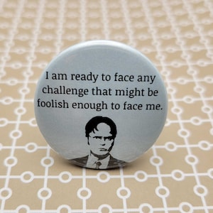 May include: A round button with a light blue background and black text. The text reads "I am ready to face any challenge that might be foolish enough to face me." A black and white image of a man with glasses is below the text.