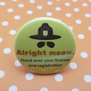 May include: A yellow button with a black and white illustration of a police officer wearing sunglasses and a hat. The text on the button reads "Alright meow. Hand over your license and registration."
