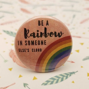 May include: A round, pink button with the text "BE A rainbow IN SOMEONE ELSE'S CLOUD" in black script. A rainbow graphic is featured below the text. The button is set against a patterned background.