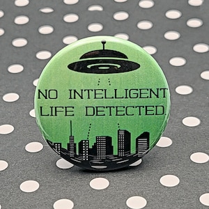 May include: A round, green button with the text "NO INTELLIGENT LIFE DETECTED" in black. The button features a black silhouette of a UFO above a city skyline. The background is a gray surface with white polka dots.