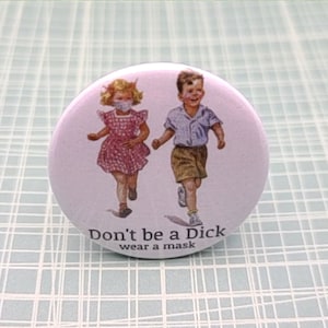 Może przedstawiać: Biały guzik z różowym tłem przedstawiający vintage'ową ilustrację dwóch biegnących dzieci. Tekst "Don't be a Dick wear a mask" jest nadrukowany na czarno u dołu guzika.