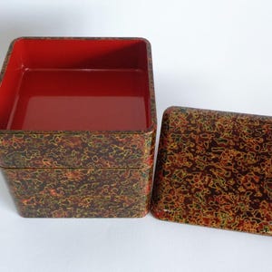 May include: A square, two-tiered stackable container with a matching lid. The exterior features a mottled, multi-colored pattern of red, yellow, and black. The interior of the container is a solid, glossy red finish with smooth, rounded corners.