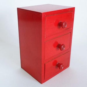 Op de afbeelding: Een kleine, rechthoekige houten opbergkast met een felrode afwerking. Deze verticale organizer is voorzien van drie gestapelde lades, elk uitgerust met een bijpassende rode ronde knop die in het midden van het voorpaneel is geplaatst.