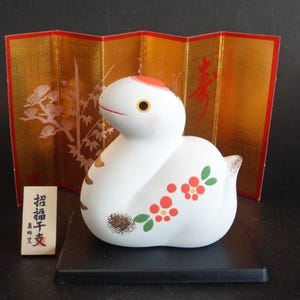 May include: A white ceramic snake figurine with a red crown, orange eyes, and floral patterns in red and green. It sits on a black base in front of a gold folding screen with red calligraphy and white pine tree illustrations, accompanied by a small wooden sign.