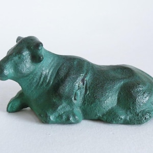 Puede incluir: Una figura de vaca de hierro fundido de color verde azulado en posición reclinada. La escultura tiene una superficie texturizada y está detallada con definición muscular visible. La vaca es un artículo decorativo, adecuado para la decoración del hogar.