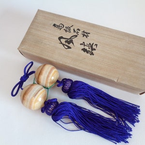 May include: Two beige and brown striped spheres with blue tassels attached to a brown box with Japanese characters on the top. The box is labelled "本場 鍛錬球".