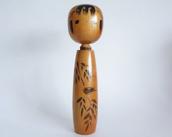 May include: A traditional Japanese wooden doll, known as a kokeshi, with a painted face and a black and brown design of a bird on a branch. The doll is made of wood and has a round head and a long, slender body.