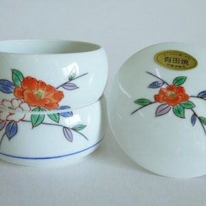 Op de afbeelding: Een witte keramische kom met een deksel, beide versierd met een bloemmotief met rode, oranje en witte bloemen met groene bladeren. Het deksel heeft een gouden label met de tekst "有田焼" in het Japans.