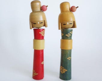 May include: Two wooden Kokeshi dolls featuring light-colored heads with painted facial features and small pink spheres attached to the side. One doll has a red body with yellow patterns, and the other has a dark green body with yellow patterns.