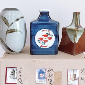 Pode incluir: Três vasos de cerâmica de várias formas e cores. O vaso da esquerda é oval com fundo cinza claro e desenhos de folhas marrons. O vaso do meio é azul com um círculo branco e um desenho floral vermelho. O vaso da direita é marrom com uma parte superior verde.