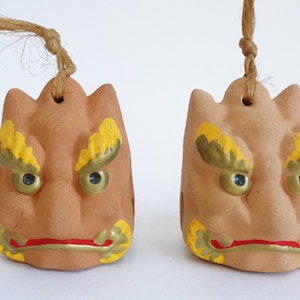 Op de afbeelding: Twee terracotta-kleurige, gestileerde gezichtsornamenten met gele en gouden accenten. De gezichten hebben rode monden, zwarte ogen en een lus om op te hangen. Elk ornament is bevestigd aan een natuurlijke vezelkoord.
