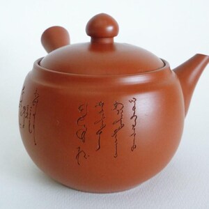 May include: A round, terracotta-colored clay teapot featuring a side handle, a short spout, and a lid with a rounded knob. The exterior surface of the teapot is decorated with vertical lines of dark, calligraphic script etched into the clay body.