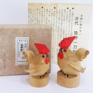 Könnte beinhalten: Zwei hölzerne Hahnenfiguren mit roten Kämmen und Kehllappen, die auf runden Sockeln stehen. Eine braune Pappschachtel und ein Blatt japanischer Text befinden sich im Hintergrund. Die Hähne sind mit detaillierten Federn geschnitzt.