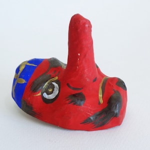 May include: A red, blue, and gold painted ceramic mask with a large, pointed nose. The mask has a black mustache and eyebrows, and a white eye with a black pupil.