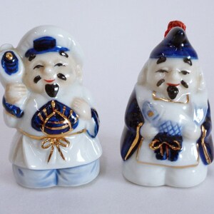 May include: Two porcelain figurines of men wearing blue and white clothing with gold accents. The figurines are holding objects in their hands.