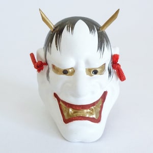 May include: A white Japanese Noh mask with black hair, gold accents, and red details. The mask features gold horns, eyes, and a red mouth. Red cords are tied on the sides. This decorative mask is a traditional Japanese art form.