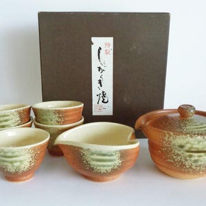 Pode incluir: Um conjunto de chá em cerâmica composto por um bule com tampa, uma taça de servir e quatro chávenas pequenas. Cada peça tem uma base castanho-avermelhada com um toque de vidrado verde texturado. Atrás encontra-se uma caixa de arrumação castanha.