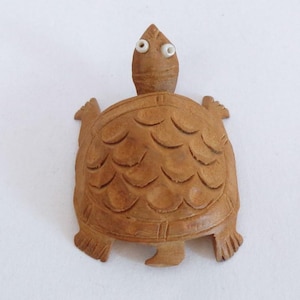 Op de afbeelding: Houten schildpadbeeldje met gesneden details. De schildpad heeft een gedetailleerd schild, een kleine kop met twee witte ogen en vier vinachtige poten. Het beeldje is gemaakt van lichtbruin hout.