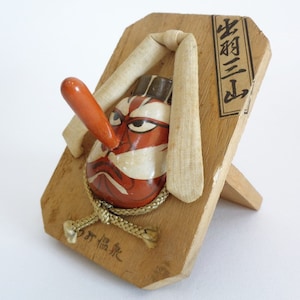 May include: A wooden Japanese geta (traditional Japanese sandal) with a painted mask of a demon with a large red nose. The geta is on a stand and has the text "寿胡三山" written on it.