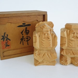 May include: Two wooden figurines of Japanese deities in a wooden box. The box has Japanese characters on the front that read "Lucky God".