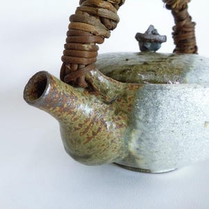 Può includere: Una teiera in ceramica con una superficie ruvida e maculata nei toni del verde oliva e del bianco sporco. La teiera presenta un beccuccio corto e largo e un coperchio con un piccolo pomello. Un manico spesso in vite intrecciata marrone è fissato.