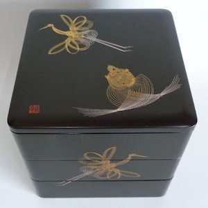 May include: A square, three-tiered black lacquer box featuring gold-coloured line art illustrations of cranes and a turtle on the lid and sides. A small red square seal with white characters is visible on the top left corner of the lid.