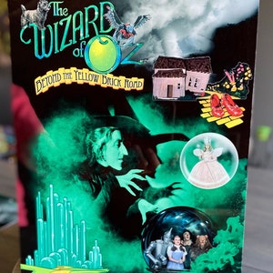 May include: A black and green metal sign with the words "The Wizard of Oz" in a green and white font. The sign also includes the text "Beyond the Yellow Brick Road" and images of the Wicked Witch of the West, Dorothy, the Scarecrow, the Tin Man, and the Cowardly Lion.