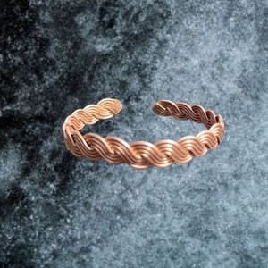 Puede incluir: Un brazalete de cobre con un diseño trenzado. El brazalete está hecho de múltiples hilos de alambre de cobre que se tejen juntos para crear una apariencia texturizada, similar a una cuerda.