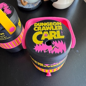 May include: Black and yellow Dungeon Crawler Carl container with pink detailing. The container features a pink handle and the text "Dungeon Crawler Carl" in yellow and pink. The container is cylindrical with a yellow and pink striped design.