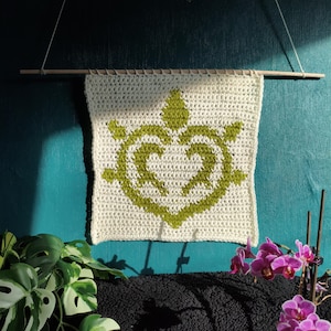 Op de afbeelding: Een crèmekleurige, gehaakte wandhanger met een groen schildpadontwerp. De schildpad staat centraal op het vierkante stuk en is opgehangen aan een houten deuvel met touw. De achtergrond is een turquoise muur met planten en bloemen.