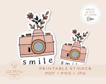 Può includere: Adesivo stampabile con due macchine fotografiche rosa con fiori in cima. La macchina fotografica più grande ha la parola "smile" scritta sotto. La macchina fotografica più piccola ha la parola "smile" parzialmente visibile. Il testo "PRINTABLE STICKER PDF I PNG I JPG" è nella parte inferiore dell'immagine.