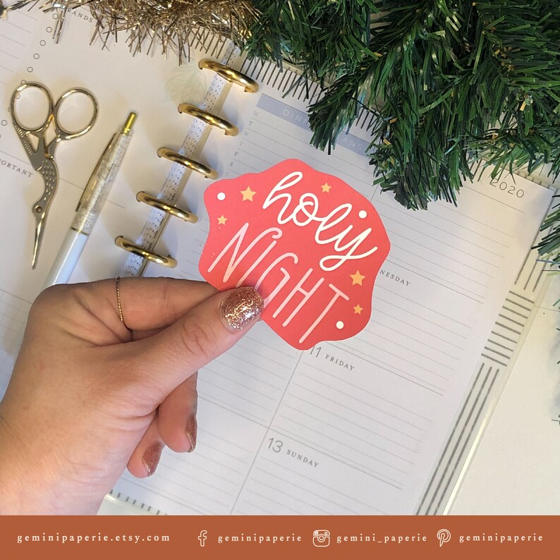 May include: A pink sticker with the text "holy night" in white cursive lettering and gold stars. The sticker is on a white planner with gold rings.