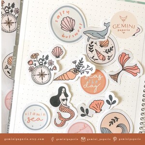 May include: A collection of stickers featuring a variety of ocean-themed designs, including seashells, mermaids, a compass, and the phrase "seas the day". The stickers are in a pastel color palette with pink, blue, and green accents.