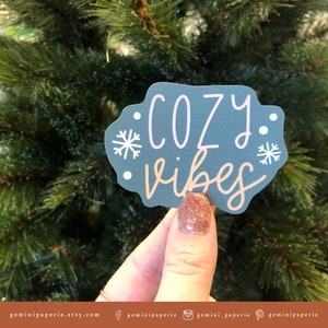 May include: A gray sticker with the text "cozy vibes" in pink and brown lettering. The sticker has two white snowflakes on either side of the text.