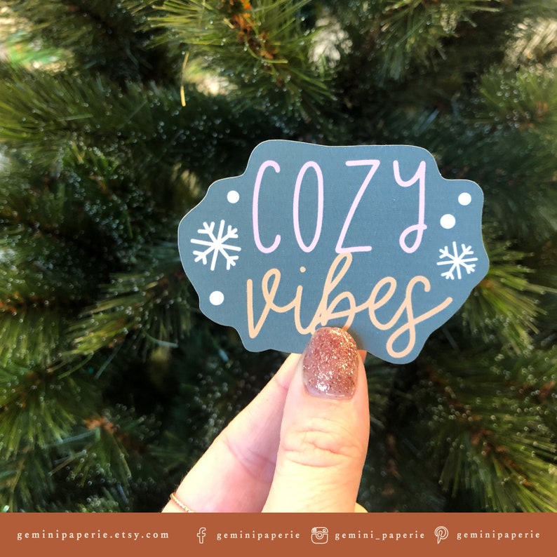May include: A gray sticker with the text "cozy vibes" in pink and brown lettering. The sticker has two white snowflakes on either side of the text.