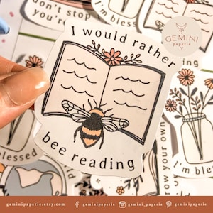 以下が含まれることがあります： 蜂と開いた本が描かれたステッカーで、テキストは「I would rather bee reading」と、花柄のデザインが施されています。ステッカーは白地に黒のアウトラインで、ピンクと黄色のカラーリングです。