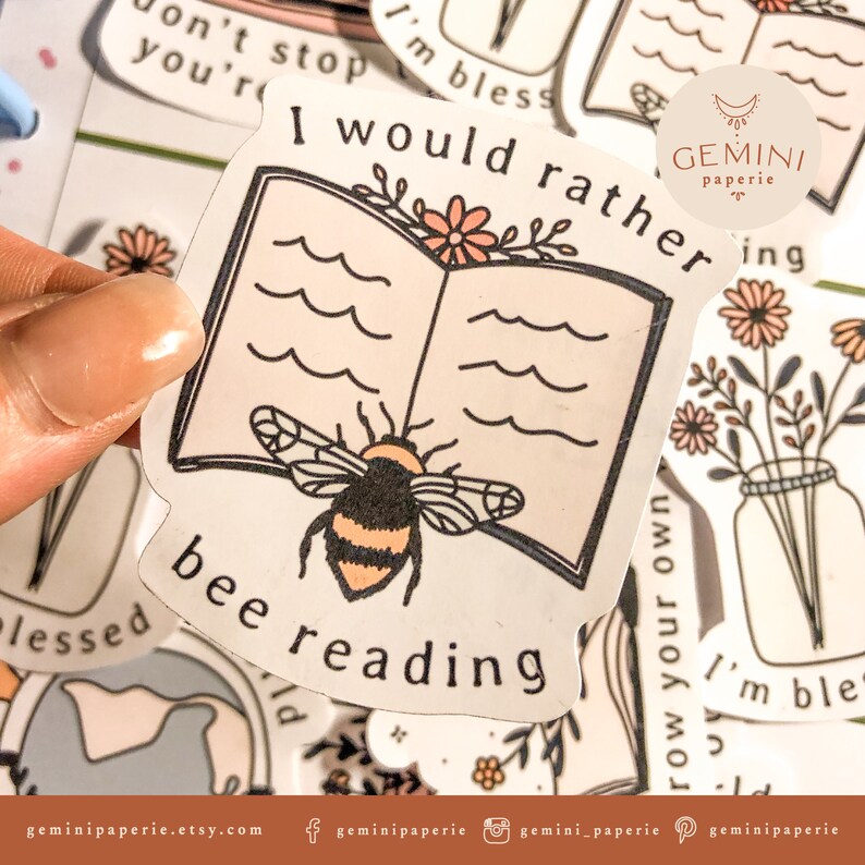 以下が含まれることがあります： 蜂と開いた本が描かれたステッカーで、テキストは「I would rather bee reading」と、花柄のデザインが施されています。ステッカーは白地に黒のアウトラインで、ピンクと黄色のカラーリングです。