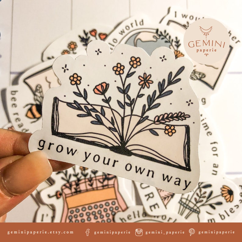 以下が含まれることがあります： 開いた本から花が咲いている白いステッカー。ステッカーには「grow your own way」と書かれています。